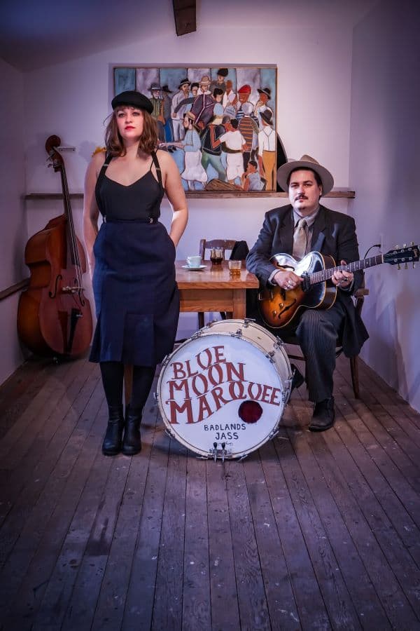 A brunette woman wearing a black dress and baret stands next to a kick drum with the group logo on the head, a man sitting to the side of the drum wearing a dark suit and camel fadora holds a hollow body guitar.