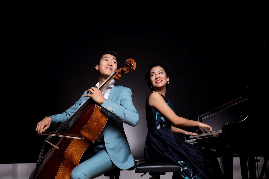 Bryan and Silvie Cheng sitting back to back, Silvie is sitting at a piano and Bryan is holding a cello