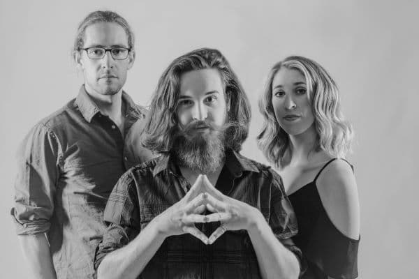 The three members of Damn tall Buildings standing in a triangle formation, the man on the left is tall and is wearing rectangular glasses, in the center a man with shoulder length hair, a long handlebar moustache and beard cover his face, on the right a blonde woman whos waivy hair sits just above the shoulders is wearing a black dress and wistfully looks on