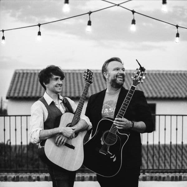 Two men stantging together looking off camera, the man on the left is younger with shaggy dark hair, on the right is a slightly older man with a short beard and his hair combed back, they both are holding acoustic guitars