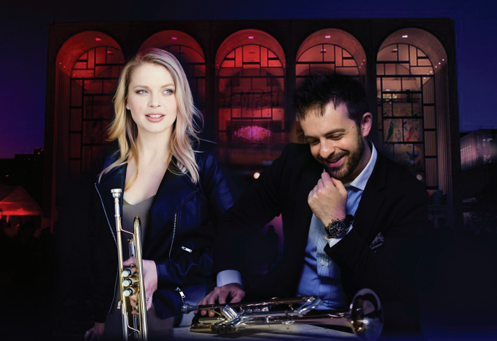 A blonde woman looks forward holding a trumpet while a man sits to her right looking down and to the right at his trumpet