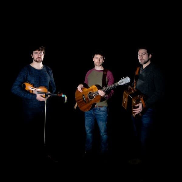 Three men stand in front of a black background, on the left a man in a blue sweater hods a violin, in the center is a man holding an acousic guitar, and on the right, almost hidden by the shadows is the accordion player holding his instrument